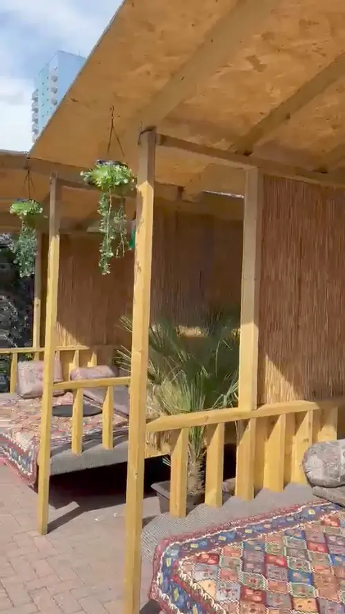 Inside one of the cabanas: a low bench spread with a Persian rug and cushions, screened with reed matting, greenery hanging from the frame.